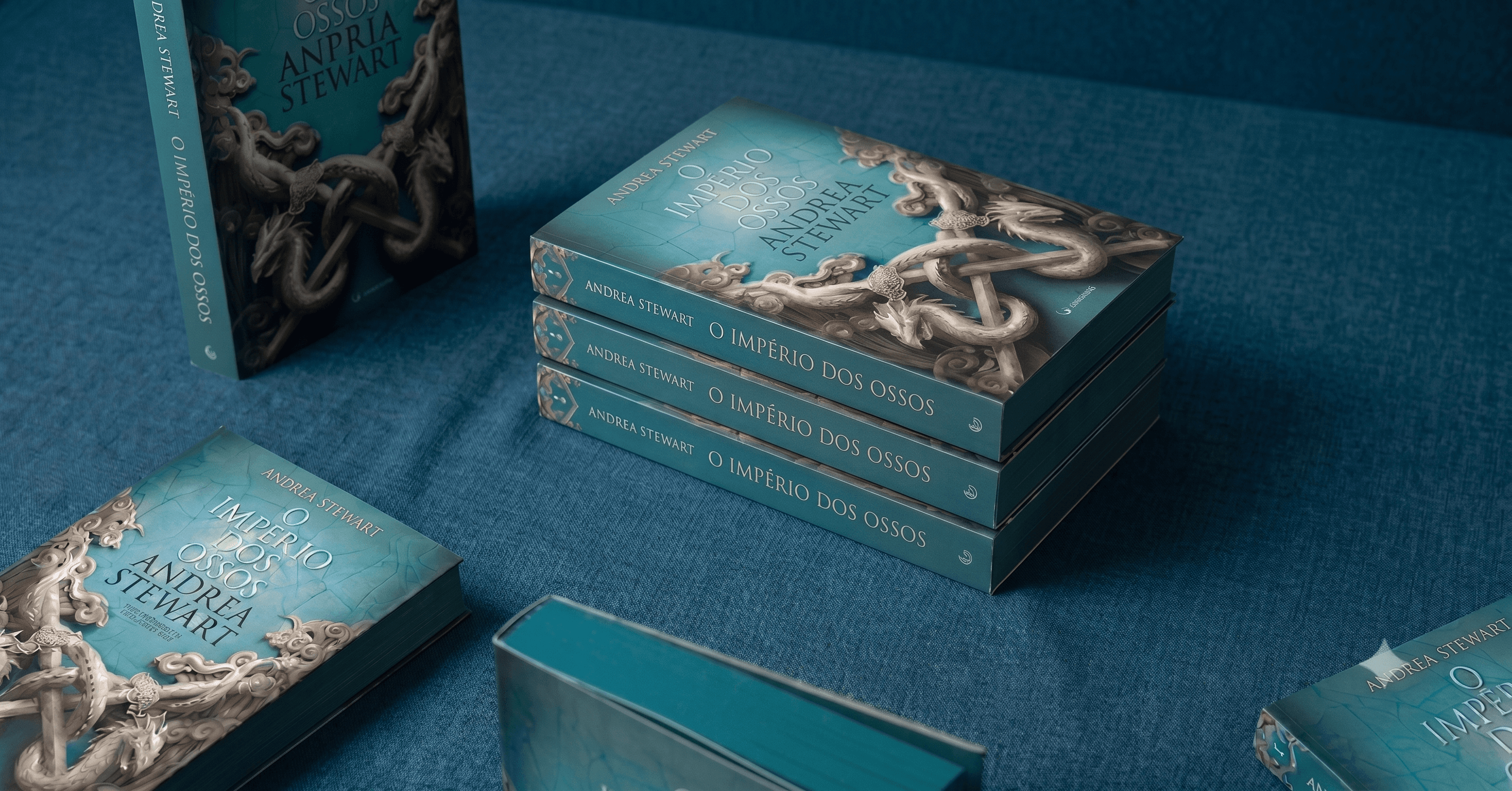 Exemplares do livro "O Império dos Ossos", de Andrea Stewart, empilhados sobre tecido azul, com as bordas das páginas pintadas de verde-água no mesmo tom da capa.